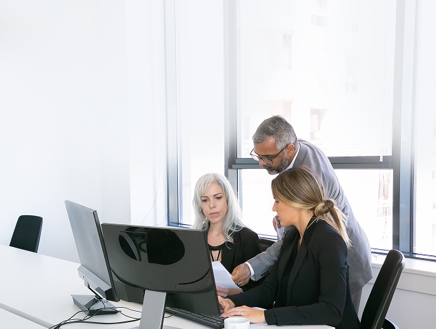 Three colleagues gathered around a desktop monitor in an office, reviewing progress on a core system offloading project